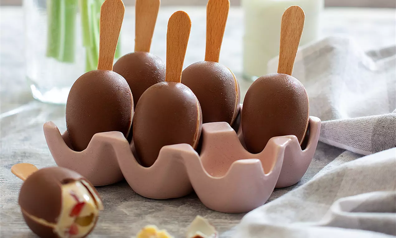 Шоколадови яйца с гладка повърхност върху дървени пръчици, подредени в розова керамична поставка за яйца, с разрязано яйце отпред