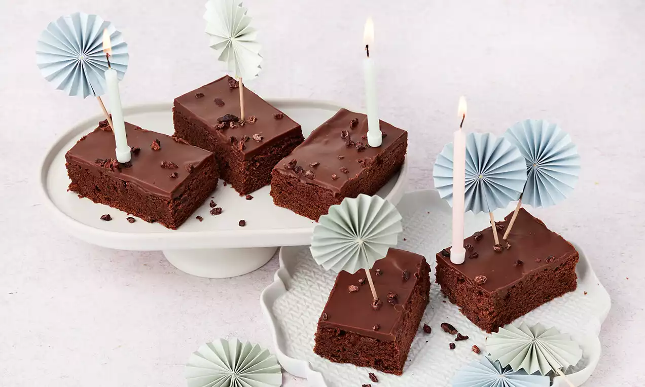 Čokoládové řezy tmavohnědé barvy s lesklou čokoládovou polevou, zdobené kakaovými boby, svíčkami a papírovými dekoracemi na bílých talířích