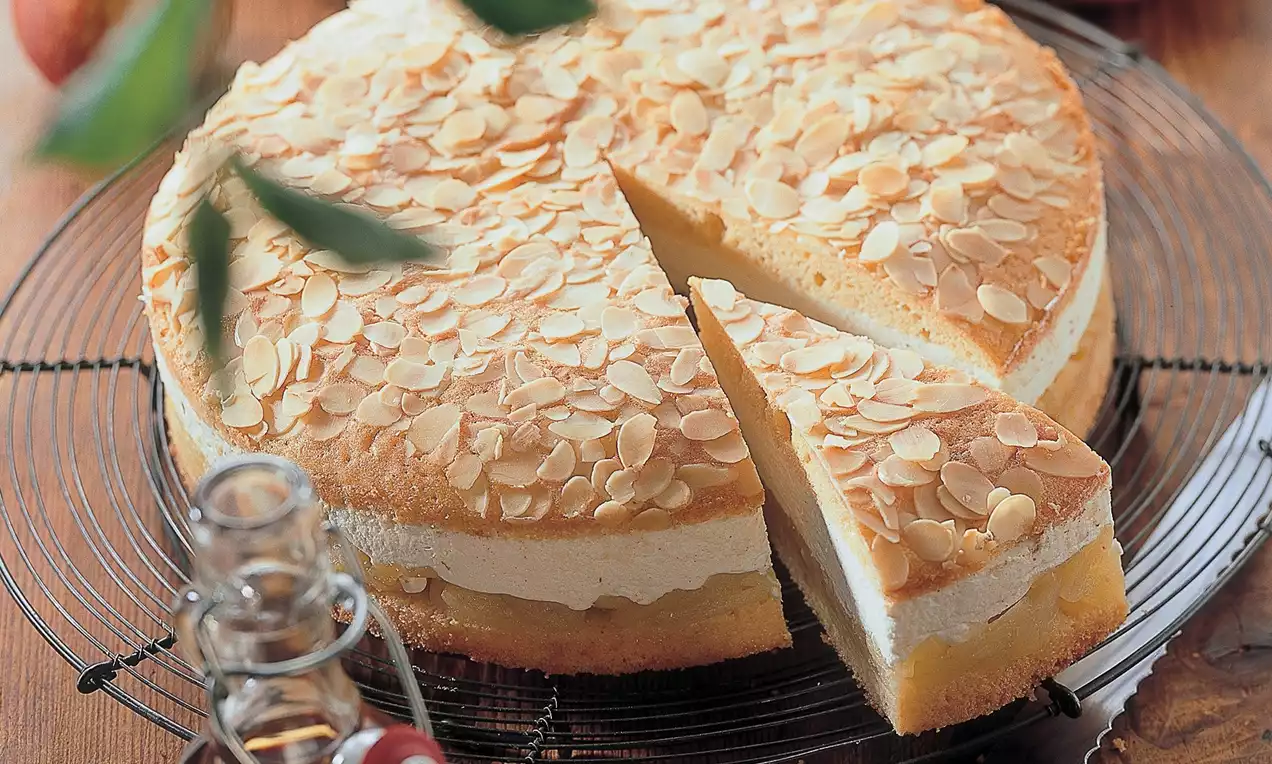 Tort rotund cu blat pufos auriu, umplutură cremoasă, presărat cu fulgi de migdale pe un grătar metalic, lângă mere și sticluța cu sirop