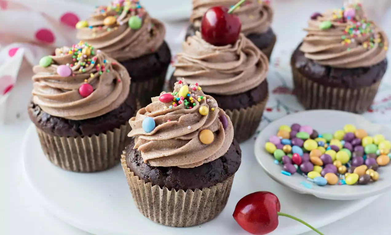 Tmavě hnědé čokoládové cupcakes s krémovou čokoládovou špičkou, zdobené barevným sypáním, třešní a podávané na bílém talíři