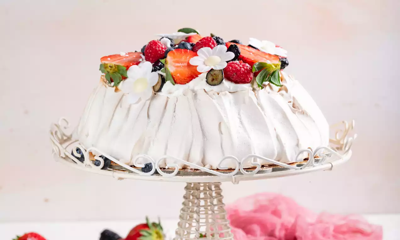 Pavlova s bijelim valovitim meringue slojem, ukrašena jagodama, borovnicama, cvjetićima i svježim listićima na bijelom stalku od metala