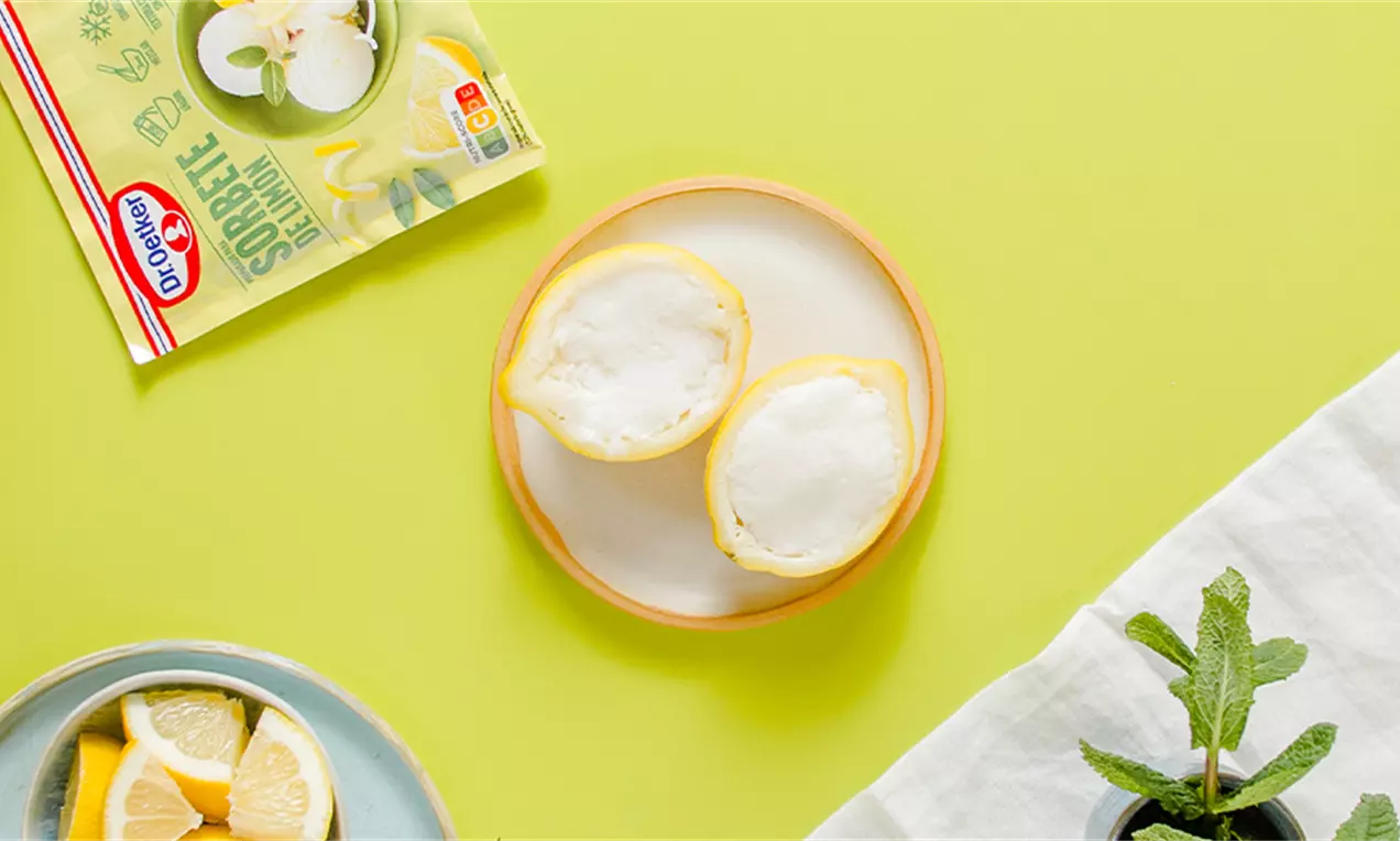 Dos mitades de cáscara de limón amarilla rellenas de sorbete blanco mate en un plato redondo, sobre fondo amarillo con decoración de menta
