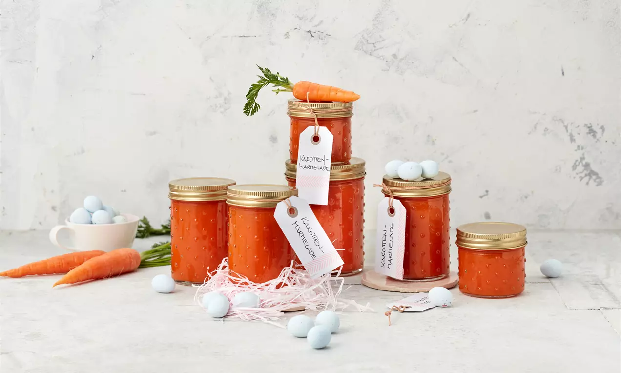 Gläser mit goldfarbenen Deckeln, gefüllt mit leuchtend orangefarbener Marmelade, dekoriert mit Namensschildern, Karotten und blauen Deko-Eiern