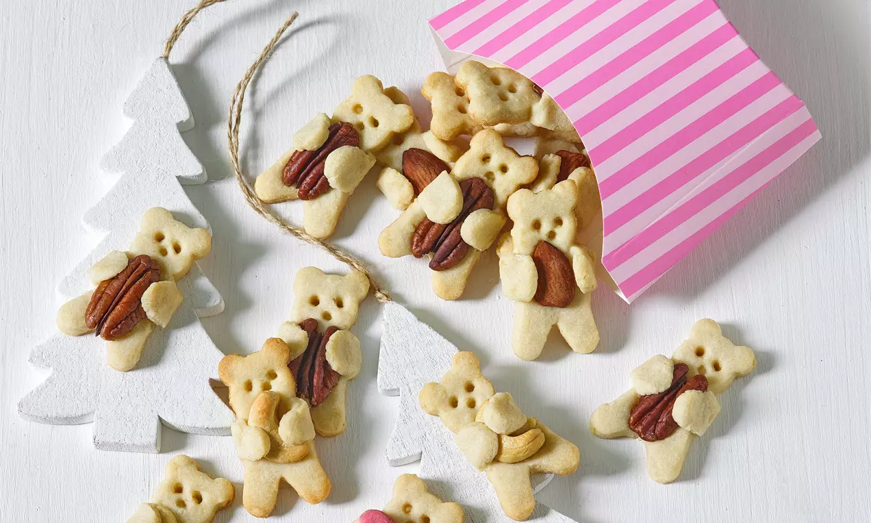 Světle béžové sušenky ve tvaru medvídků s texturou jemného těsta, každý držící oříšek, uspořádané na bílém podkladu s dekorací stromků