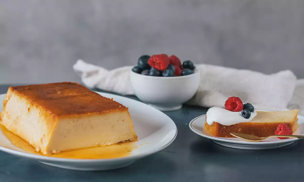 Rebanada de pudín de pan dorado y cremoso con textura suave sobre plato blanco, decorada con nata, frambuesas y arándanos
