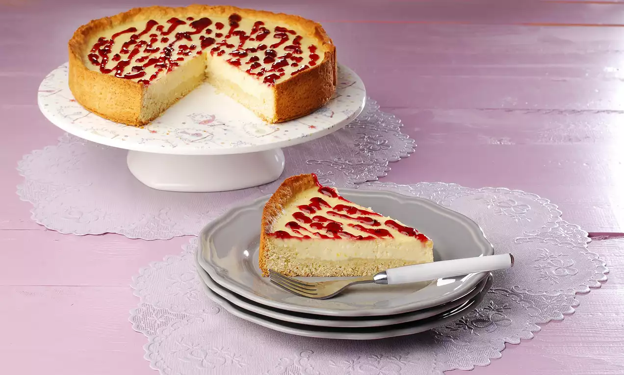 Altın rengi tabanlı lorlu cheesecake, kremamsı dolgusuyla sade beyaz tabakta vişne sosu desenleriyle zarif bir görünüm sunuyor