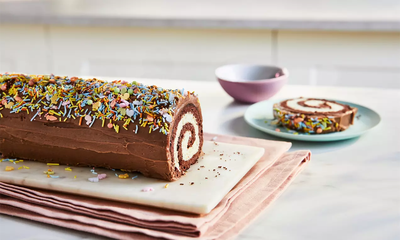 Chocolate Yule log with a spiral cream filling, smooth frosting, colourful sprinkles, served on a marble board with slices on plates