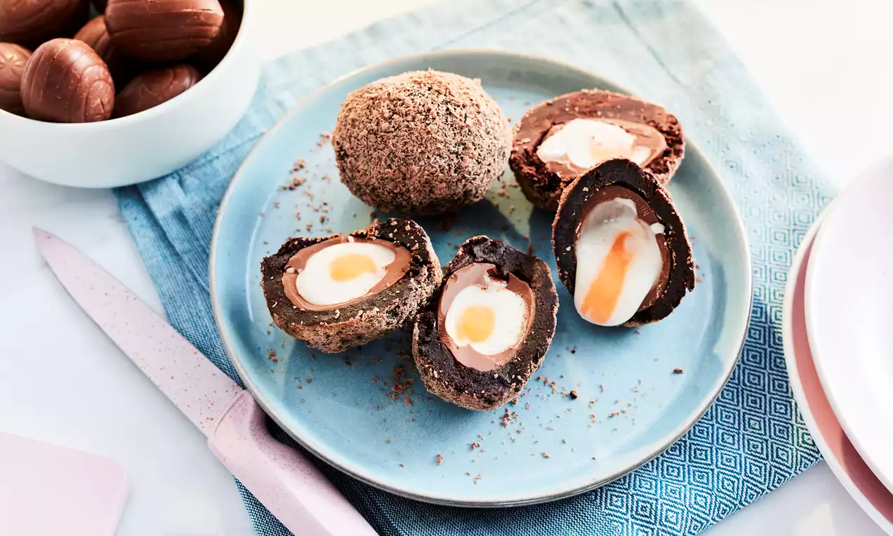 Chocolate Scotch Egg with a glossy brown shell, creamy white and orange filling, halved and whole on a pale blue plate with a textured cloth