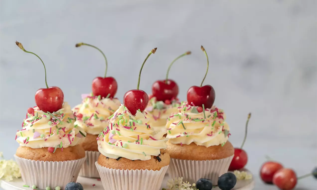 Šviesaus biskvito keksiukai su puriu gelsvu kremo sluoksniu, papuošti vyšniomis ir spalvotais pabarstukais ant šviesios lėkštės.