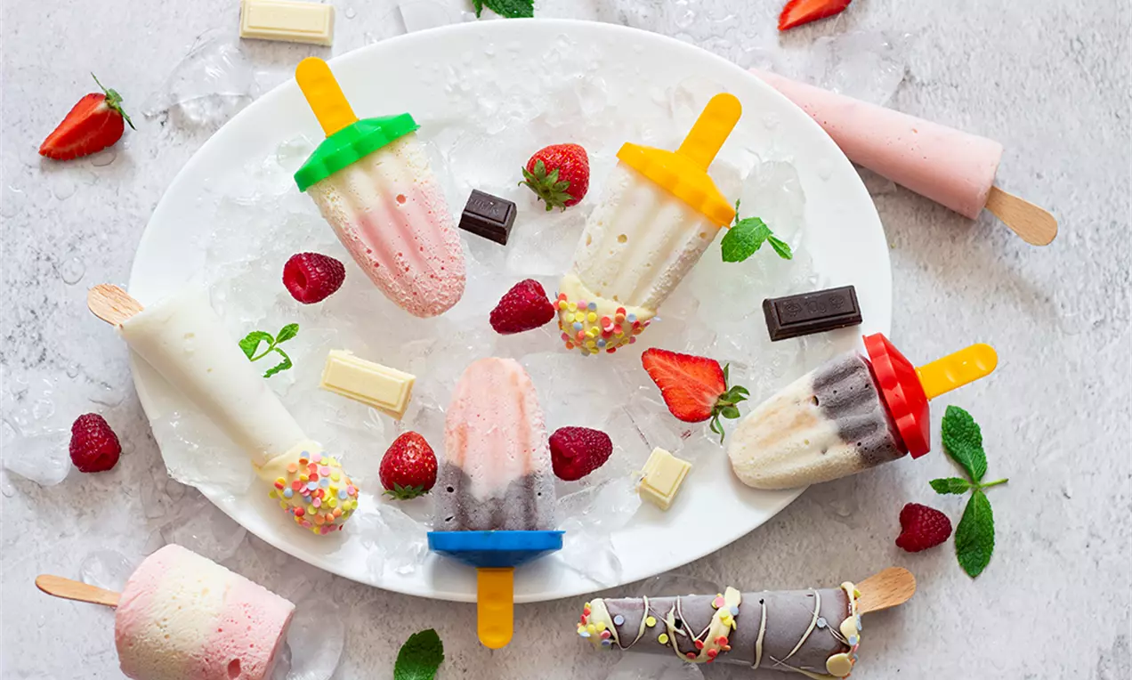Разноцветни сладоледи на дървени клечки с кремообразна текстура върху бяла чиния с лед, допълнени с ягоди, мента и блокчета шоколад