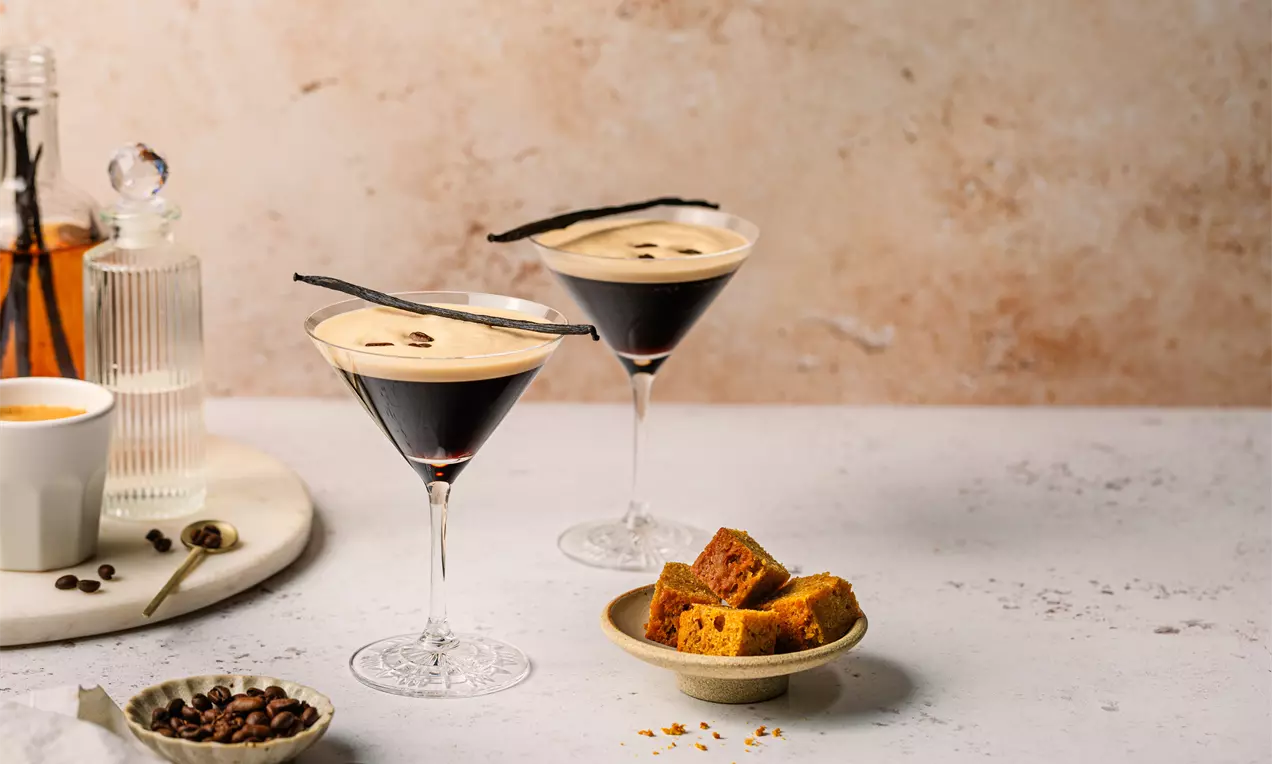 Picture - Zwei Espresso Martinis in Cocktailgläsern mit cremiger Schaumkrone und Kaffeebohnen garniert, serviert neben Würfeln von goldbraunem Kuchen