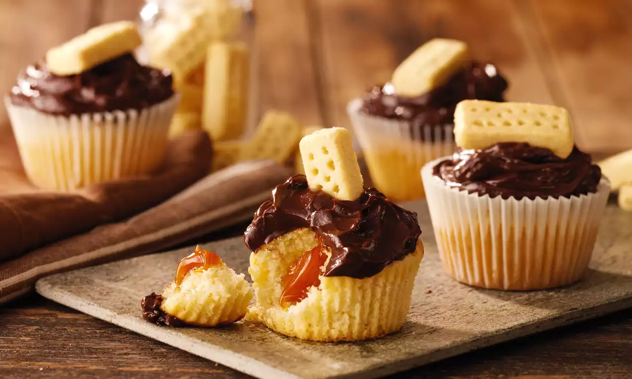 Golden muffins with crumbly shortbread biscuits, glossy chocolate topping, and caramel filling inside on a rustic wooden table