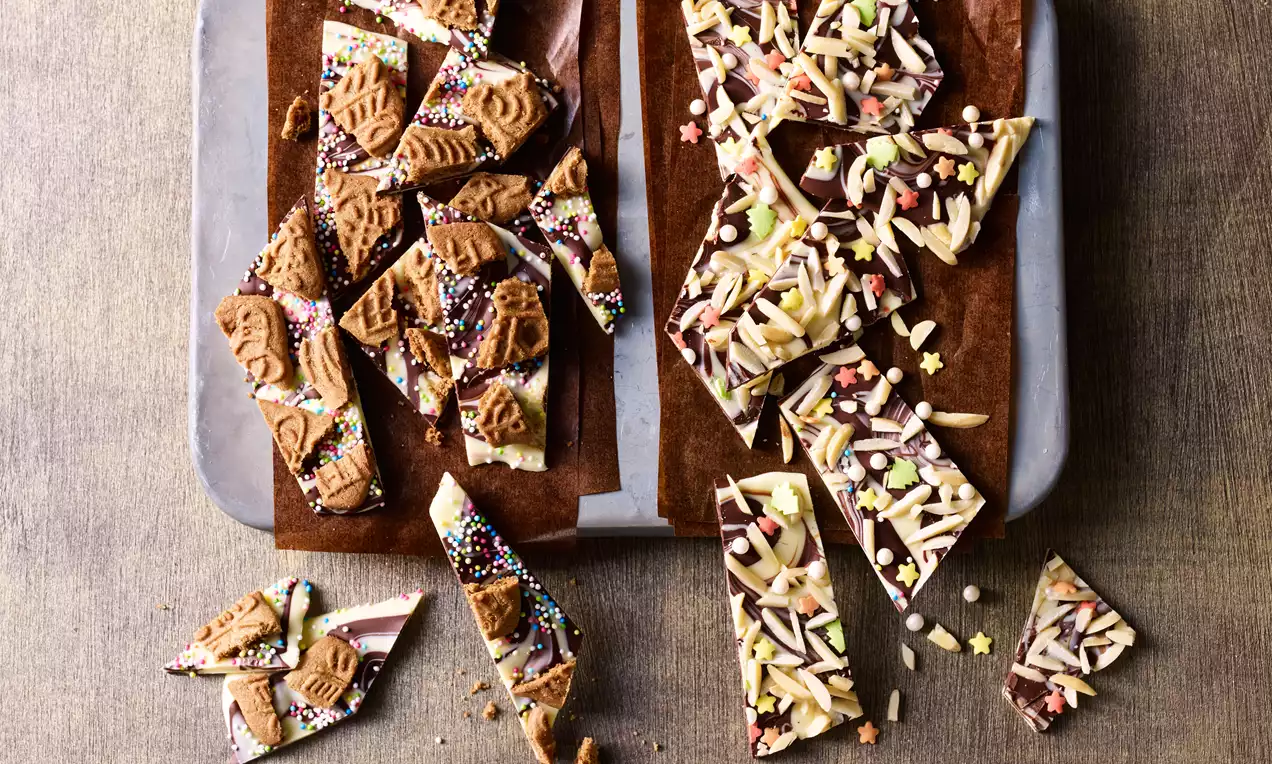 Twee soorten chocoladebark op bruine bakpapierstroken met kleurrijke sprinkles, amandelschaafsel en koekstukjes op een houten tafel