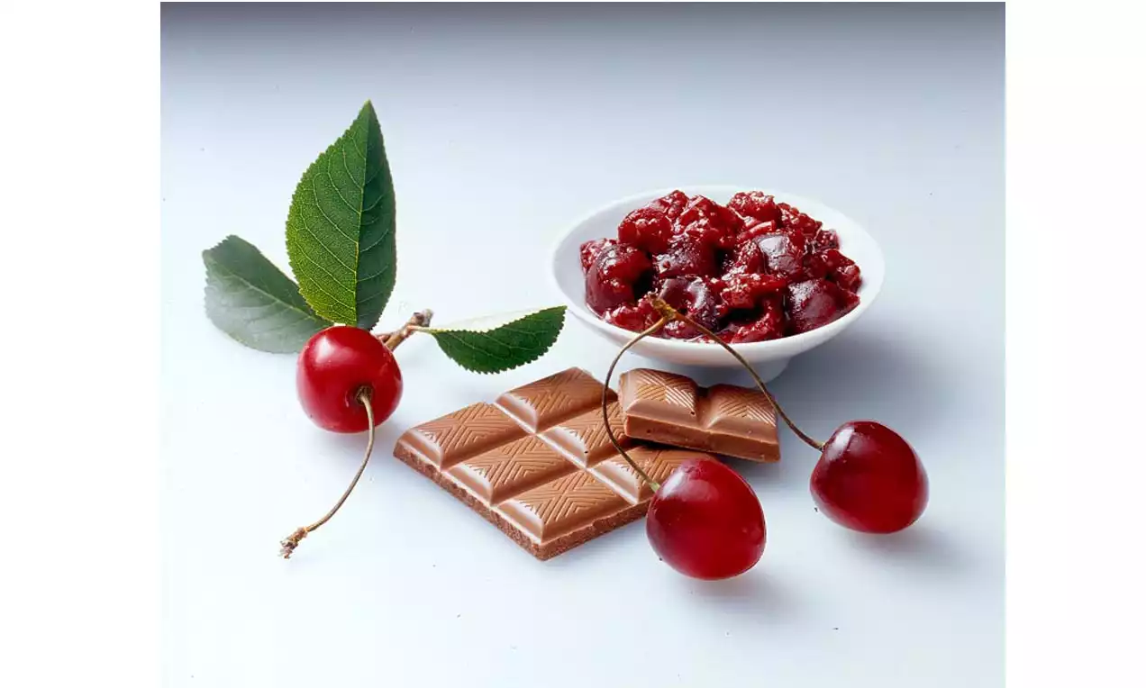 Glänzend rote Kirschen und grüne Blätter neben einem Stück brauner Schokolade und einer weißen Schale mit dunkelroter Konfitüre