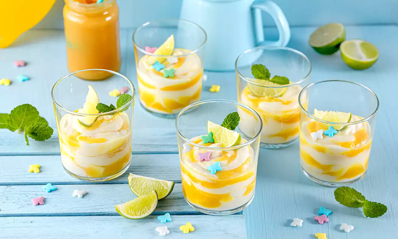 Gläser mit gelb-weißer geschichteter Creme, garniert mit Minzblättern, Limettenschnitzen und bunten Sternen auf einem hellblauen Holztisch