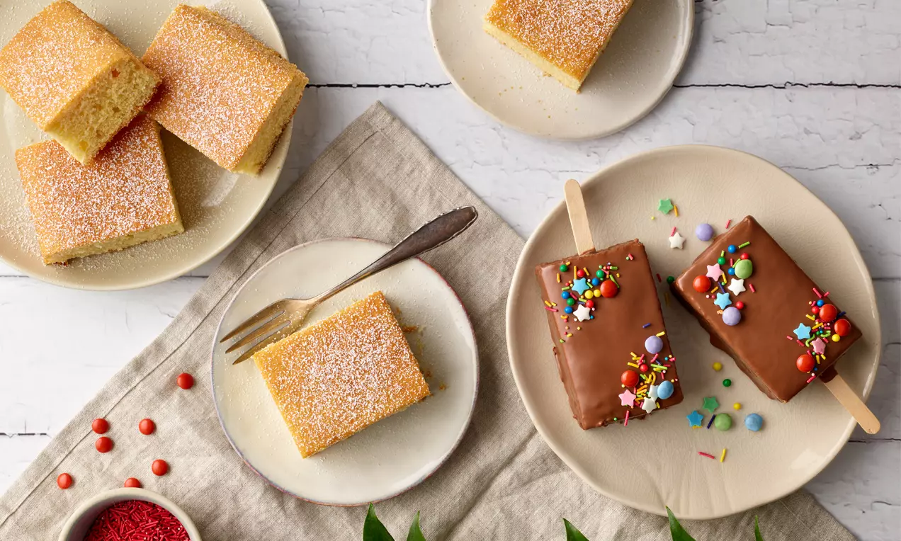 Goldgelber Blitzkuchen als rechteckige Stücke mit Puderzucker bestäubt sowie glasierte Varianten mit bunten Streuseln auf hellen Tellern