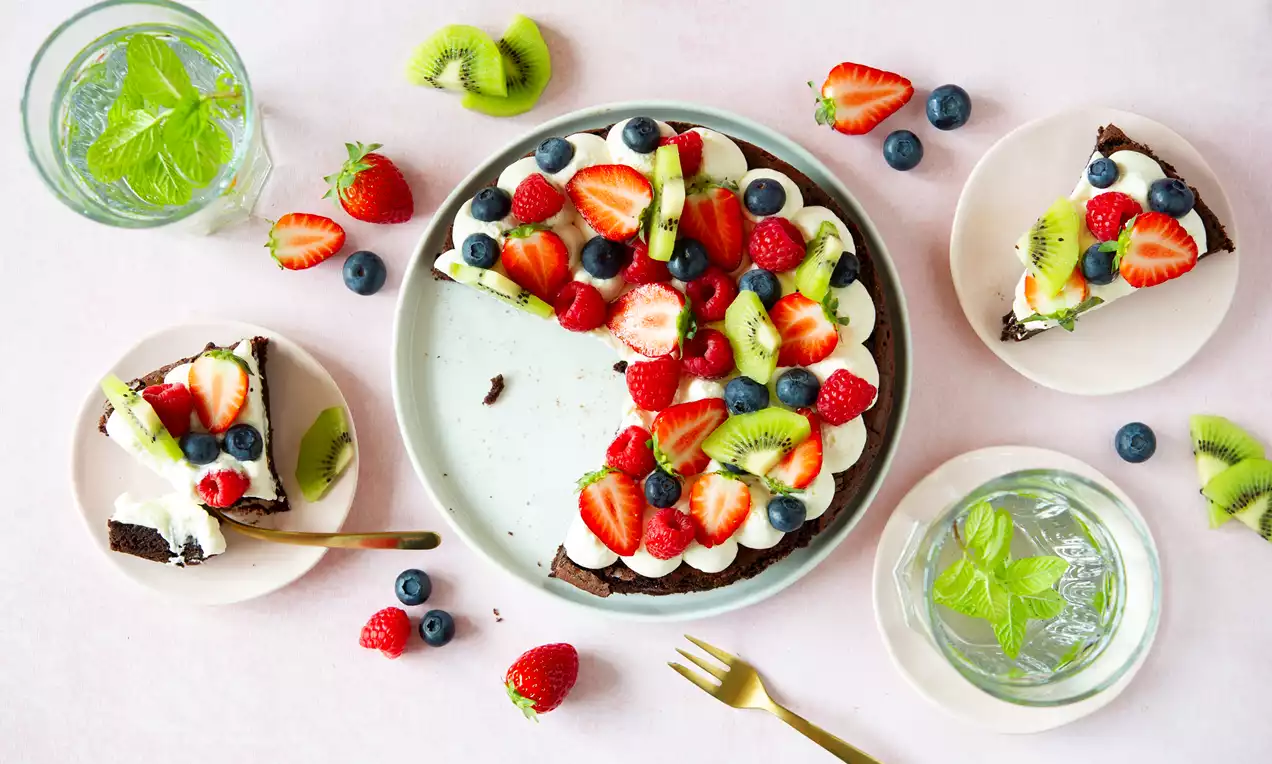Donkerbruine brownie op een licht bord, bedekt met witte room, aardbeien, kiwi, blauwe bessen en frambozen, omlijst door kleine schotels en glazen