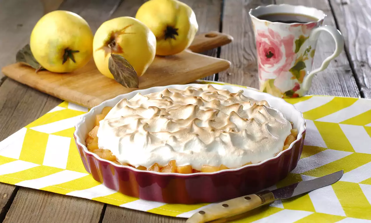 Altın rengi tart tabanı üzerinde kabarık ve hafif kızarmış bir mereng, yanında ayvalar, çiçek desenli bir fincan ve bir bıçakla sunulmuş