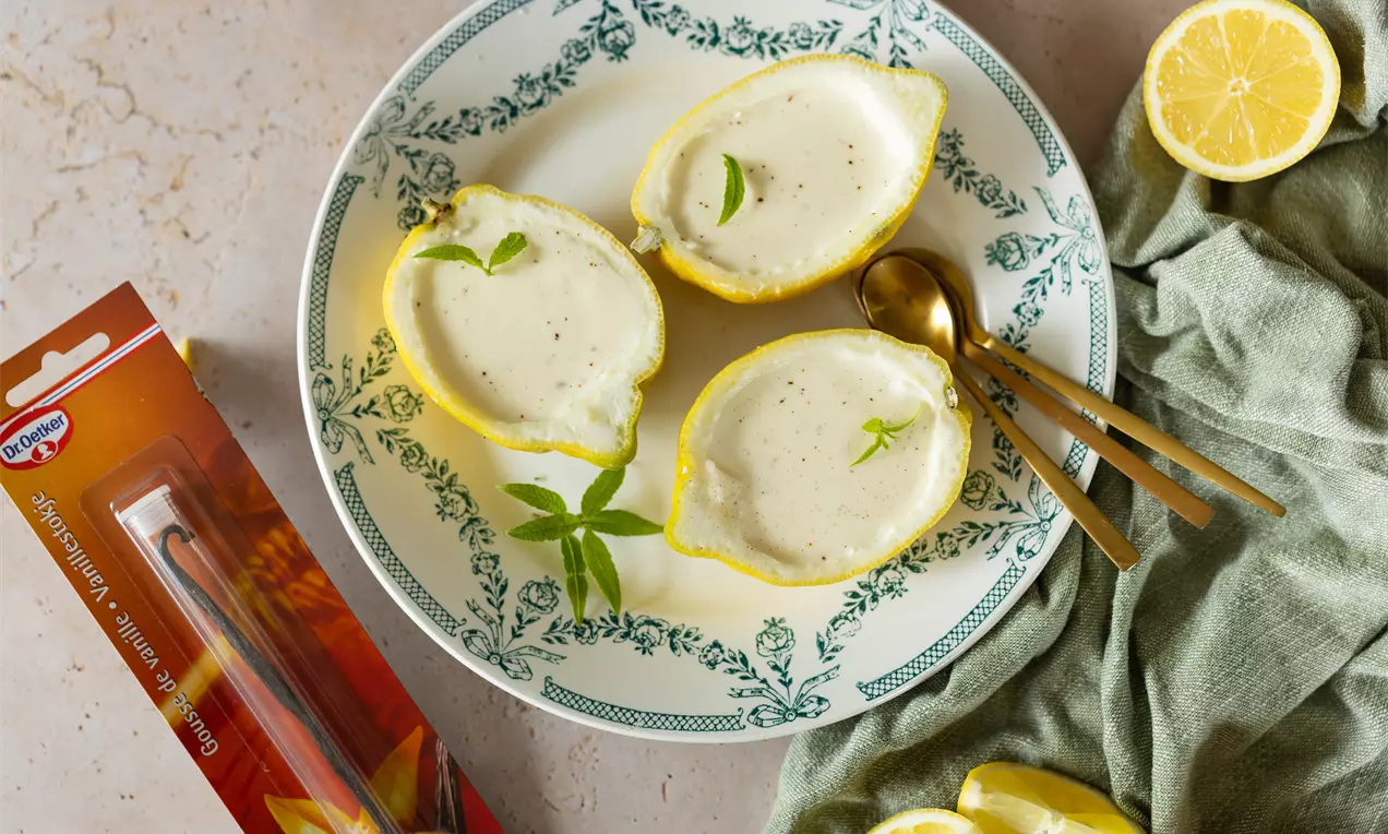 Lichtgele lemon posset met gladde, romige textuur geserveerd in uitgeholde citroenen op een wit bord met groene decoratieve rand