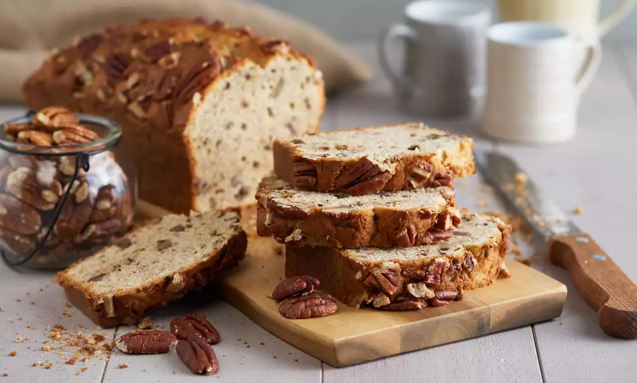 Banana bread dorato con noci pecan brunite incastonate nella superficie, tagliato a fette spesse su un tagliere di legno con gusci sparsi