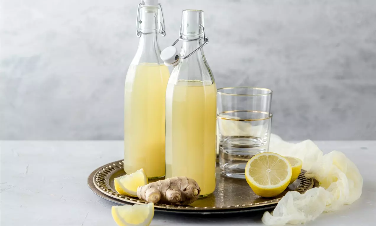 Две стъклени бутилки с бледожълт джинджифилов сироп върху метален поднос, с резени лимон, корен джинджифил и чаши на светъл фон