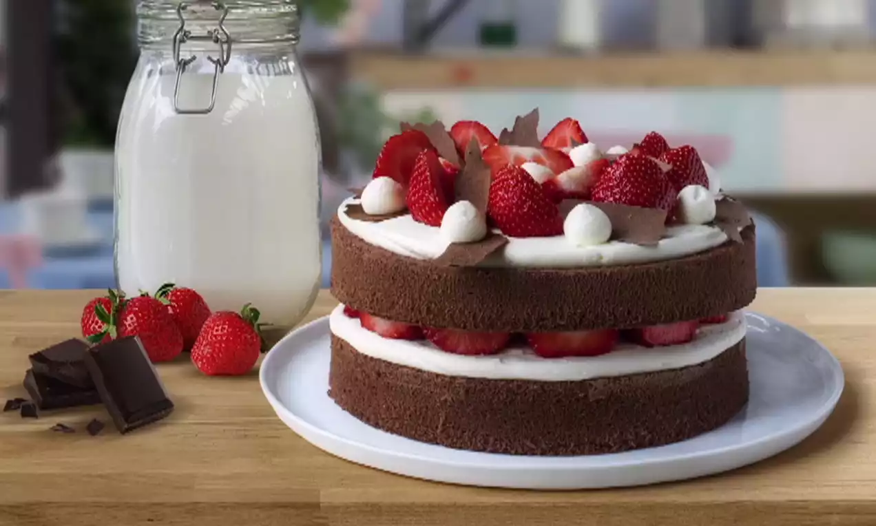 Tweedelige chocoladetaart met witte crème, verse aardbeien, chocoladeschilfers en roomtoefjes op een wit bord met melkfles en losse aardbeien