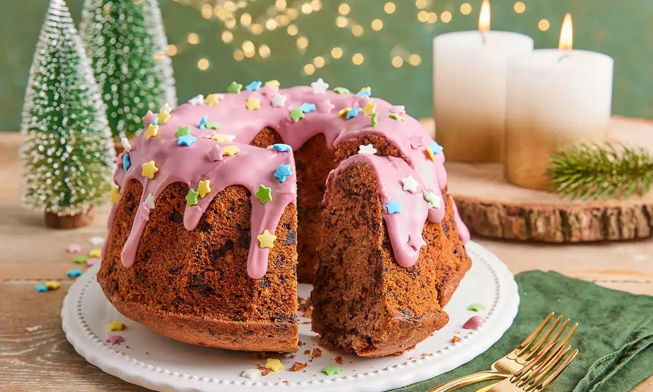 Punschkuchen mit rosa Glasur und bunten Sternstreuseln, angerichtet auf weißer Tortenplatte mit goldenen Gabeln, Kerzen und Tannenbaum-Deko