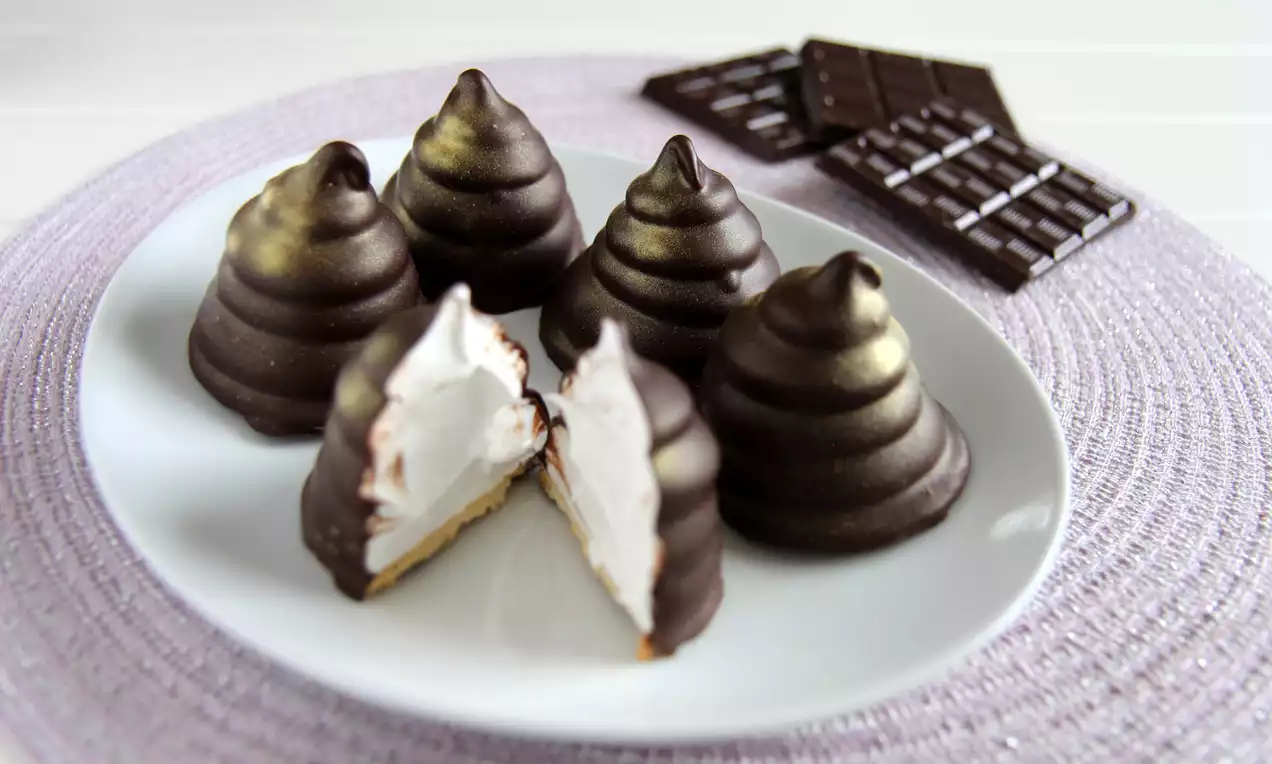 Kokosboller med mørk sjokoladeoverflate og gullspray, én delt i to med hvitt fyll, på hvit tallerken med lilla tekstil og sjokoladeplate
