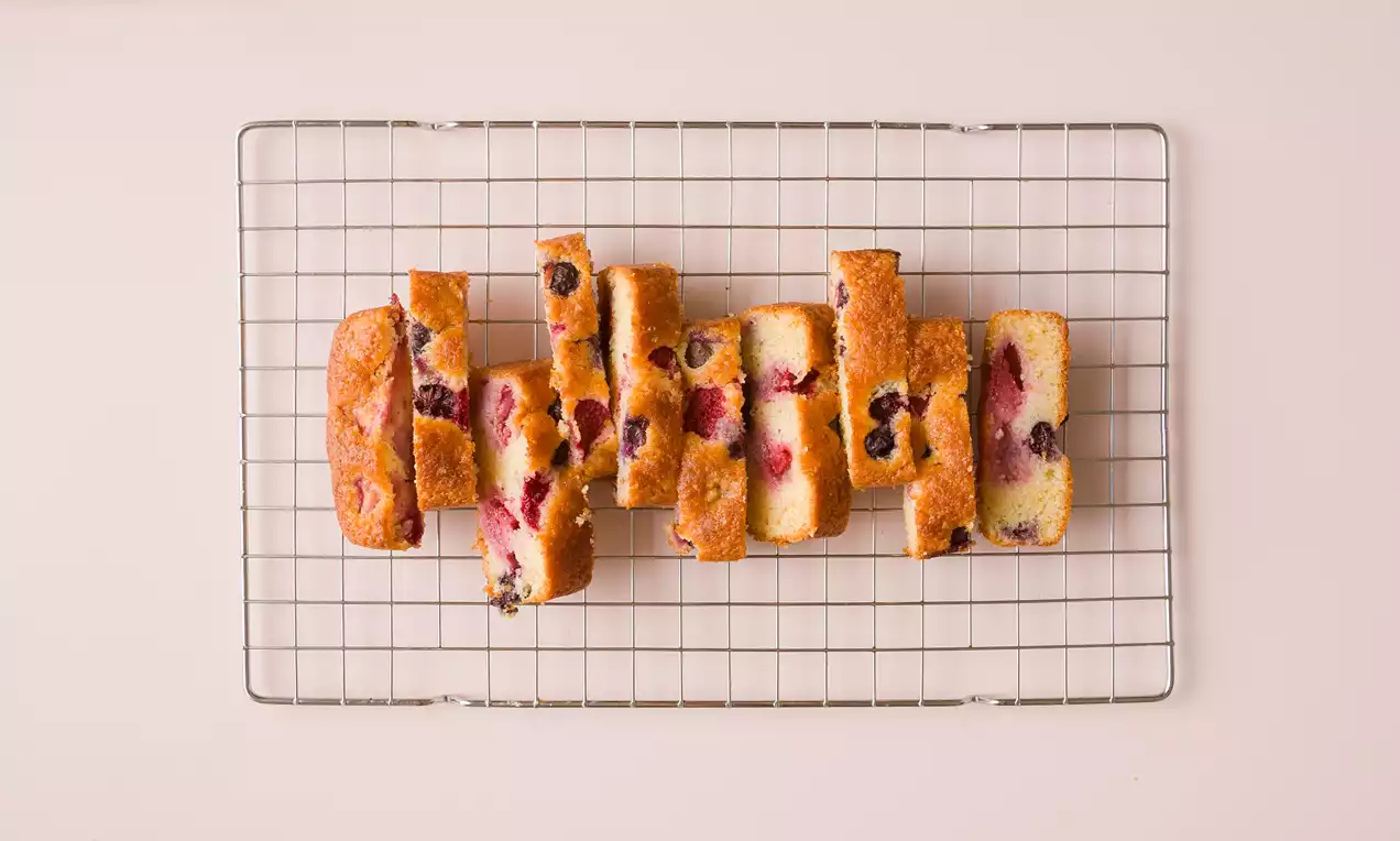 Bizcocho dorado cortado en rectángulos, con interior suave y esponjoso salpicado de frutos rojos, dispuesto sobre una rejilla metálica