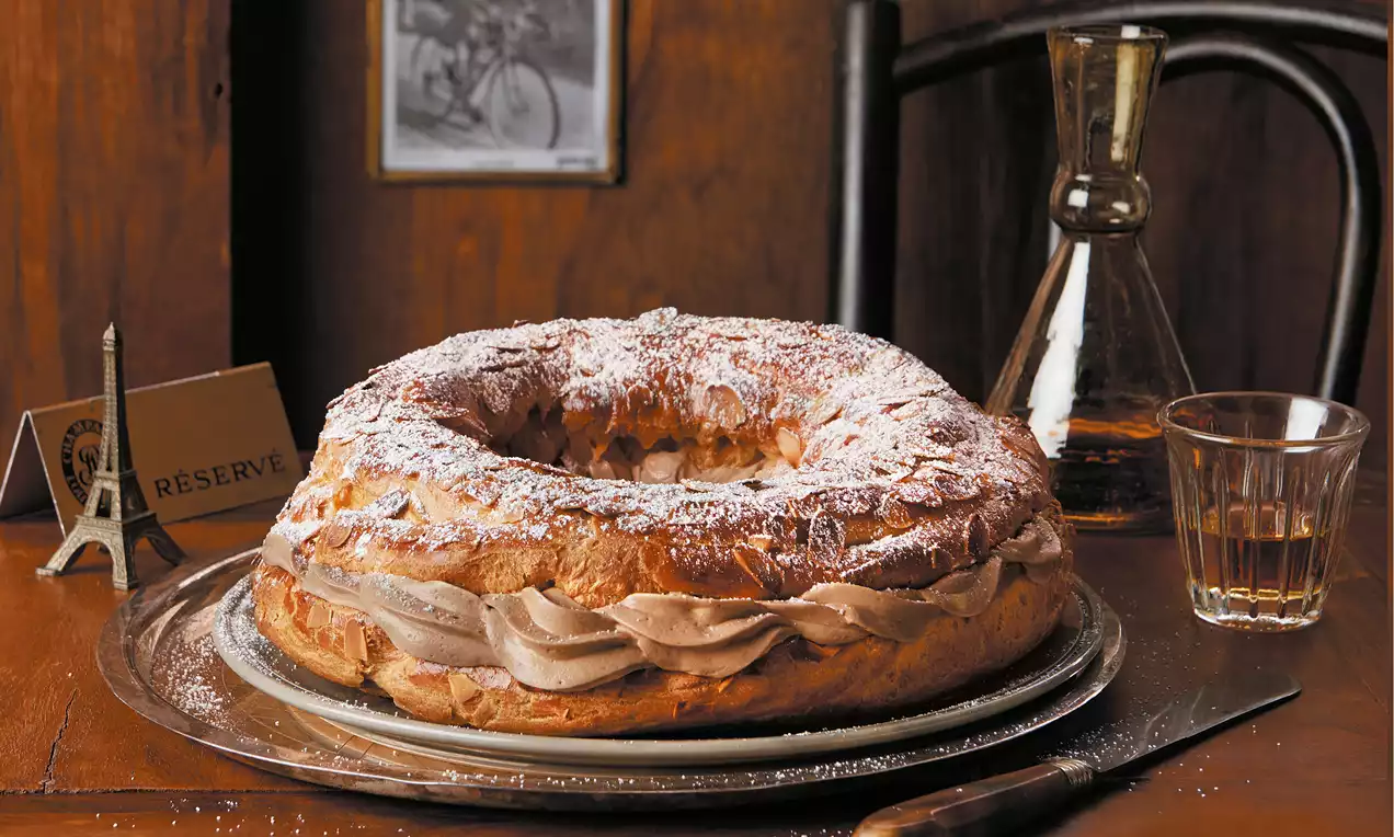 Ein goldbrauner Brandteigkranz mit sahnig-cremiger Nougatfüllung, mit Puderzucker bestäubt auf silbernem Teller, umgeben von warmem Holz-Ambiente