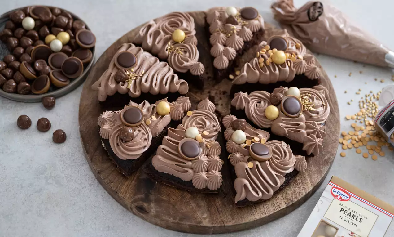 Chokoladekake delt i trekanter med brun krem, hasselnøtttopping og sjokoladekuler på en rustikk trefjøl, omgitt av konfekt og sprøytepose
