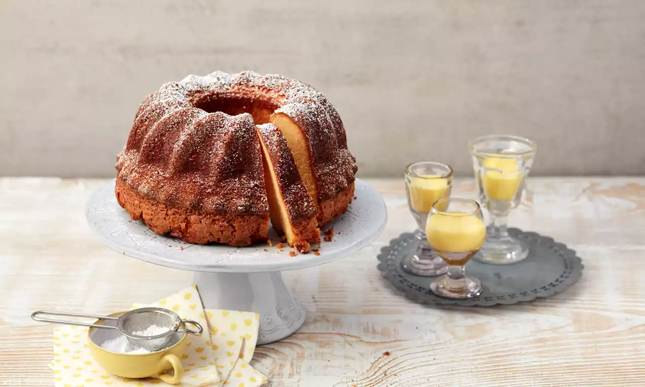 Saftiger, goldbrauner Eierlikör-Gugelhupf mit Puderzucker bestäubt auf weißer Tortenplatte, daneben Eierlikör in kleinen Gläsern