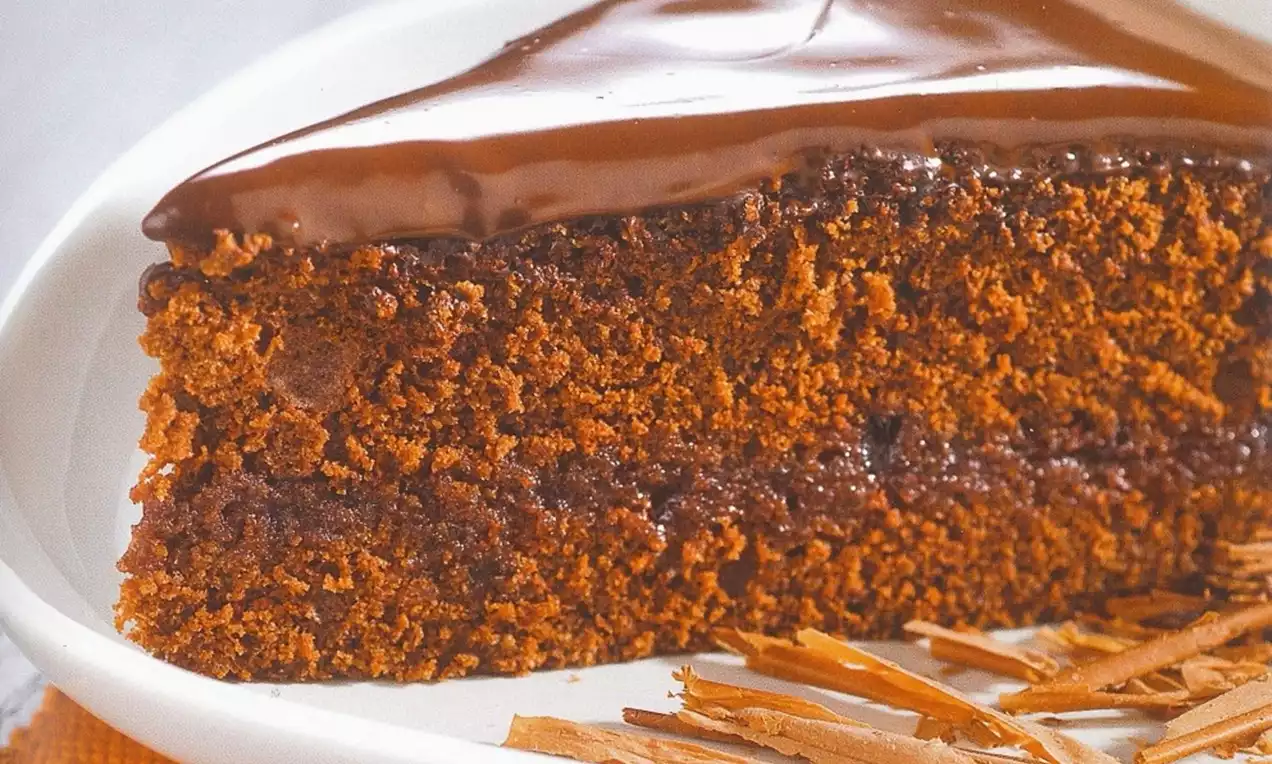 Part de gâteau Sacher brun foncé à la texture moelleuse, recouverte d'un glaçage chocolat brillant sur une assiette blanche avec des éclats de chocolat