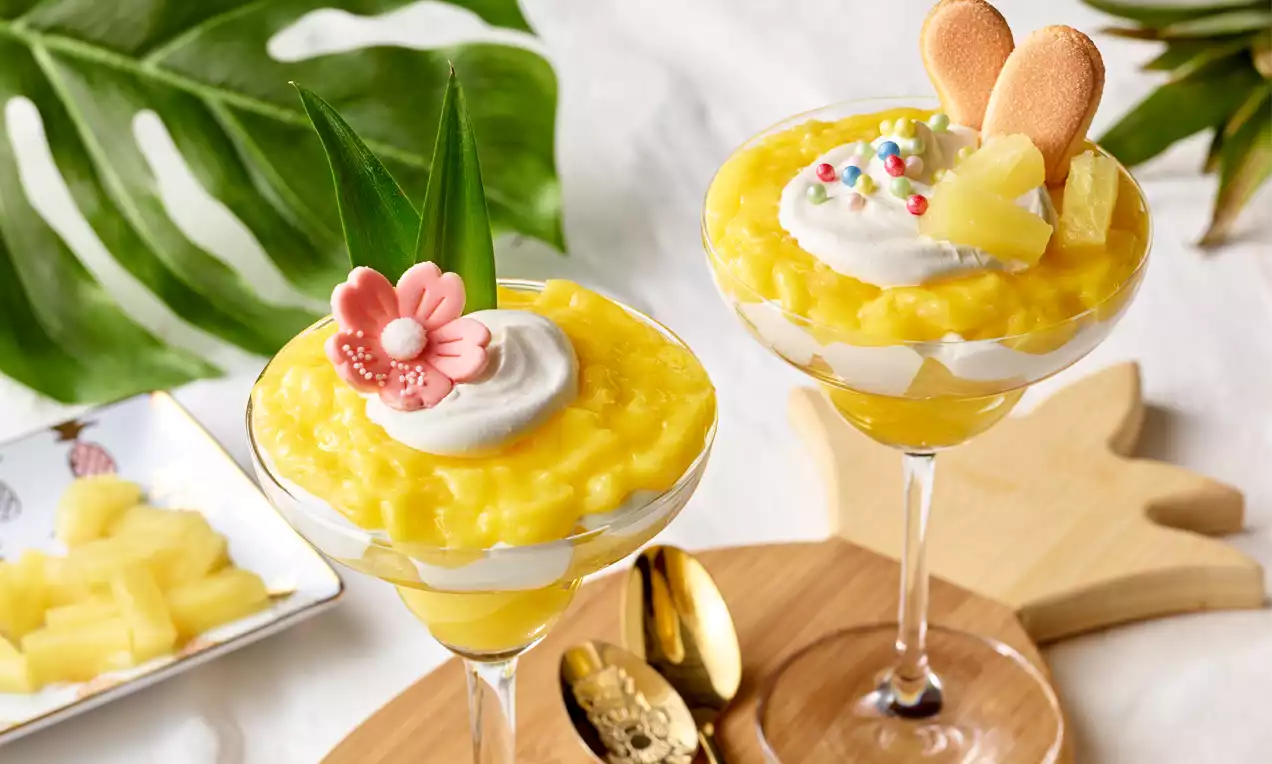 Schichtdessert aus leuchtend gelber Ananascreme und weißer Sahne in hohen Gläsern, dekoriert mit Früchten, Keksen und bunten Details