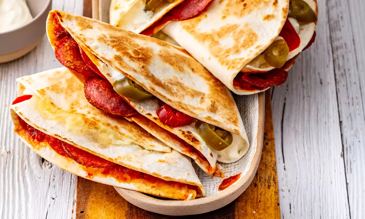 Golden pan-fried folded tortillas filled with red pepperoni, melted cheese, and green jalapeños on an oval plate on a wooden board
