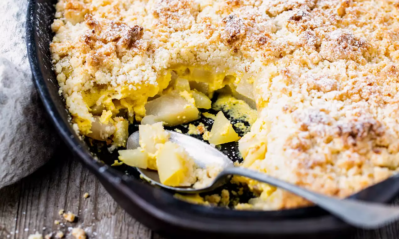 Quitten-Crumble mit goldbrauner, krümeliger Kruste über gelblichen Quittenstücken in einer dunklen Auflaufform mit silbernem Löffel