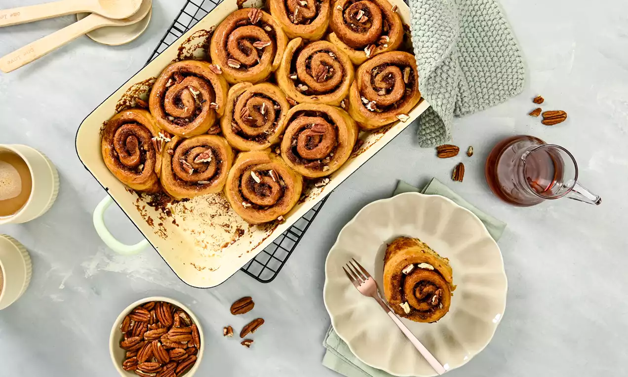 Fluffy cinnamon rolls with golden swirls, garnished with pecan pieces and syrup, on a wavy beige plate alongside scattered pecans and dishes