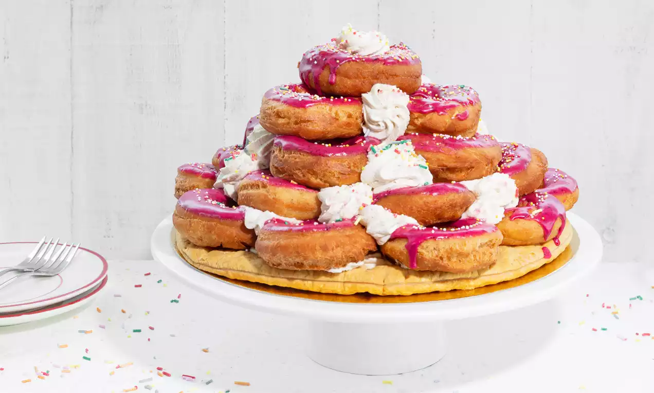 Donuttaart met lagen goudbruine donuts, roze glazuur, slagroomtoefjes en gekleurde strooisels op een wit taartplateau