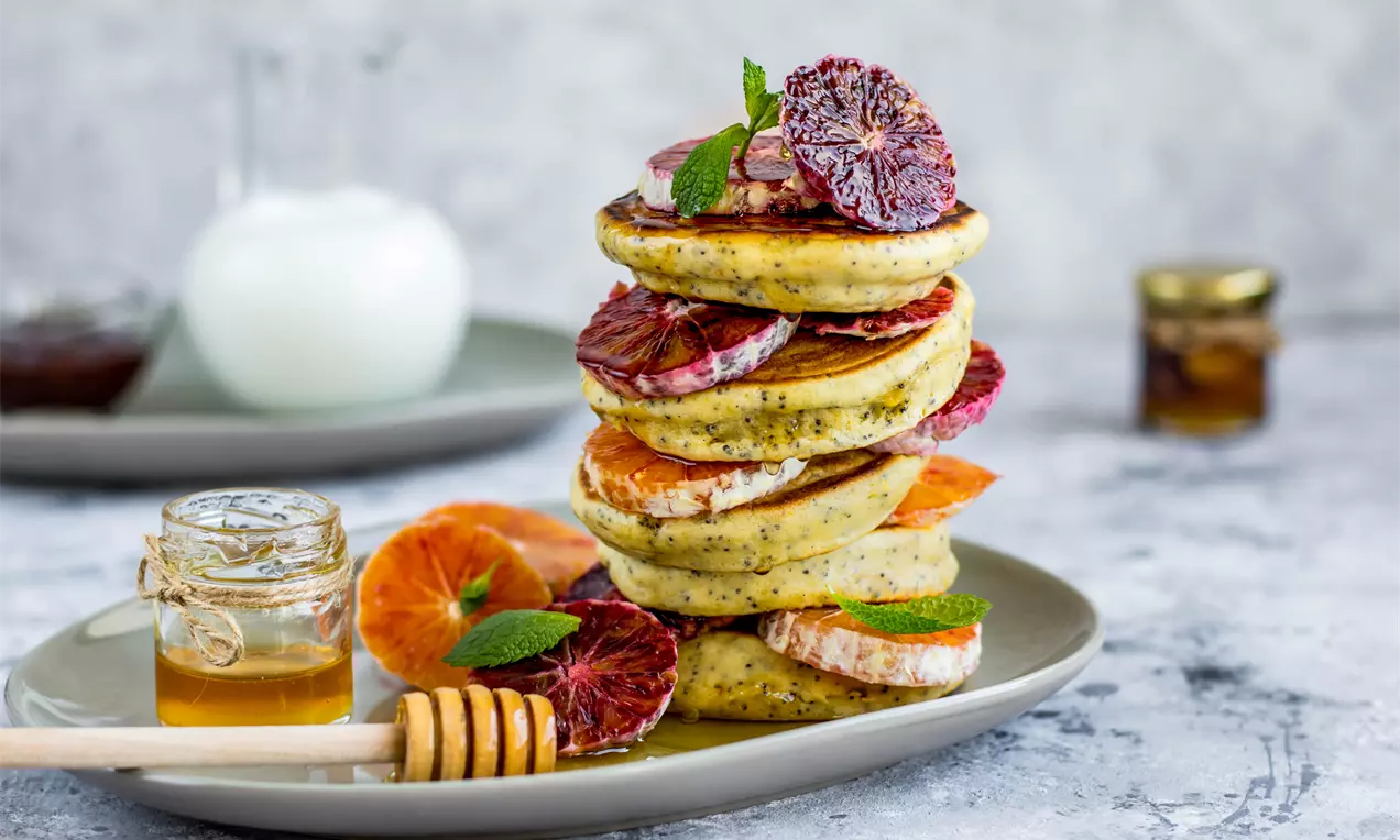 Пухкави палачинки със светъл кремав оттенък и макови точици, подредени на купчина, гарнирани с резени червен портокал и свежа мента