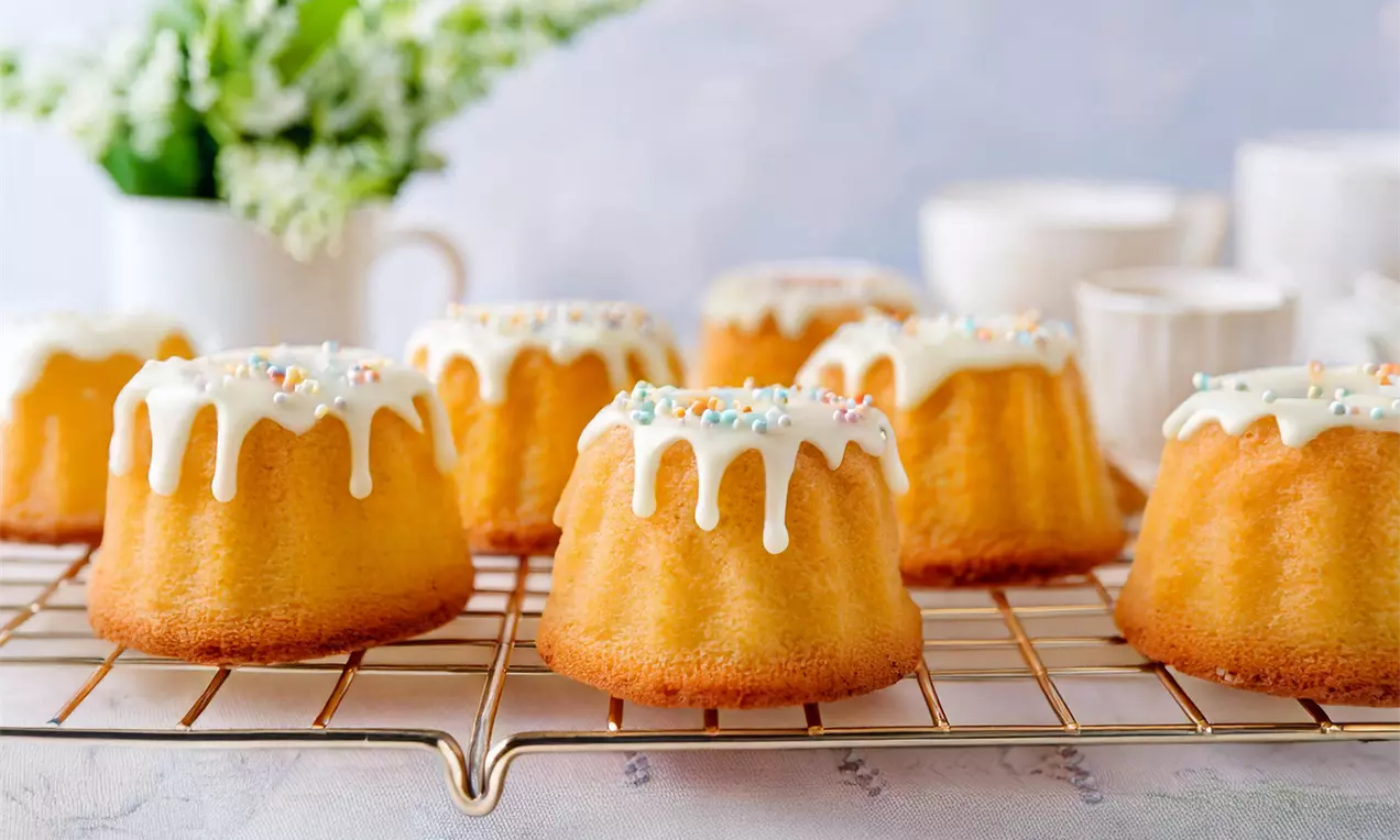 Mini kolački zlato rumene barve z belo glazuro, ki se razliva čez robove, posuti z mavričnimi sladkornimi posipi na kovinski mreži