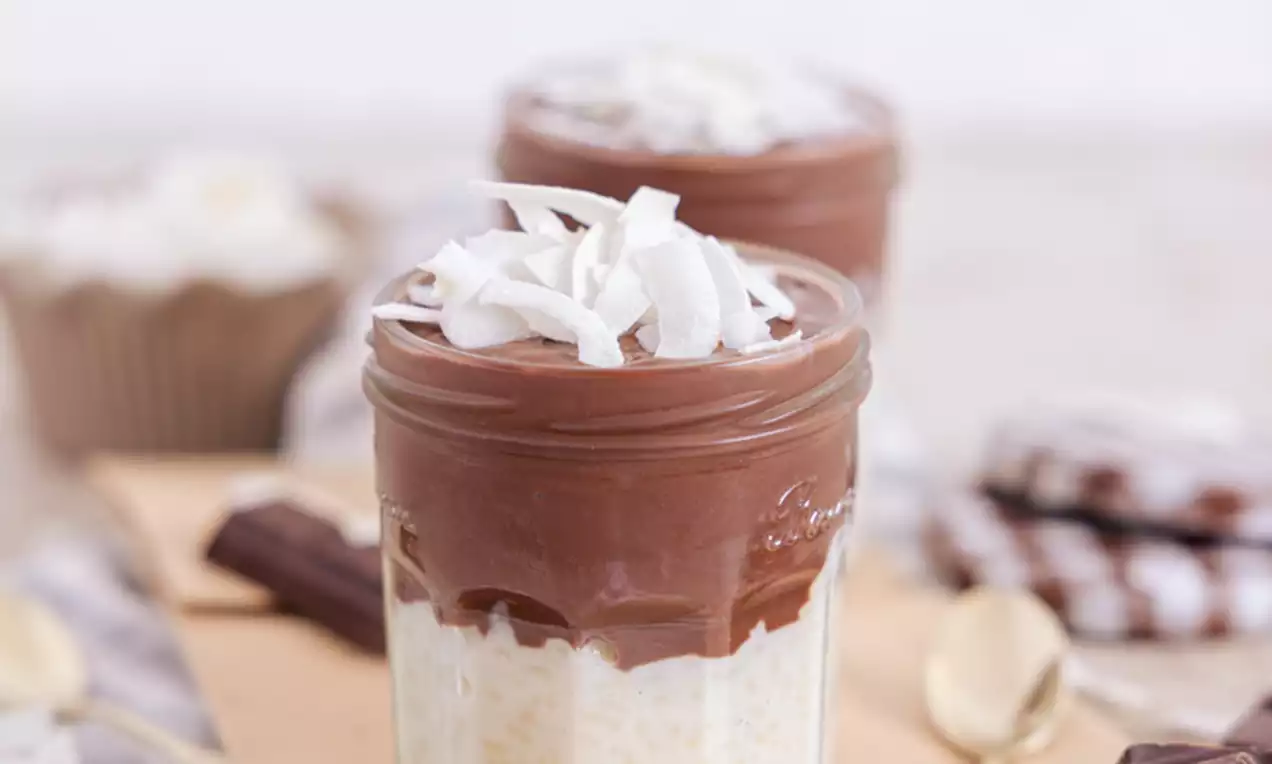 Sloj kremastega belega mlečnega riža, prekrit z gladkim temno rjavim čokoladnim pudingom in posut z belimi trakovi kokosovega čipsa v steklenem kozarcu