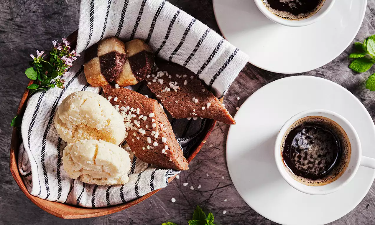 Drömmar i ljusgul ton med spröd textur ligger i en korg med randig servett tillsammans med chokladsnitt och kaffekoppar