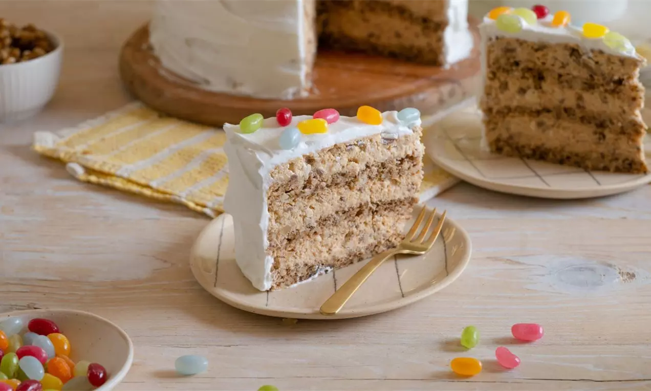 Parče kockaste torte svetlosmeđe boje s tri sloja, kremastim filom, glazurom od belog šlaga i šarenim žele bombonama na vrhu