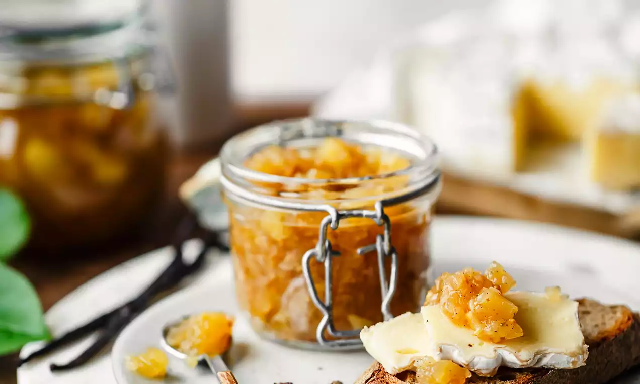 Picture - Golden apple chutney with a chunky texture in a glass jar, served on rustic bread with cheese on a white plate with scattered spices