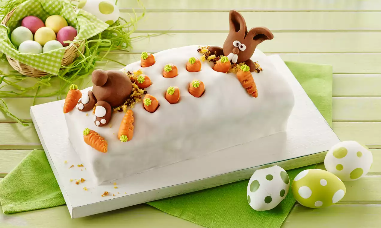 Witte, langwerpige taart met marsepeinen wortels en een chocoladekonijn dat lijkt te graven, op een wit bord met groene decoratie