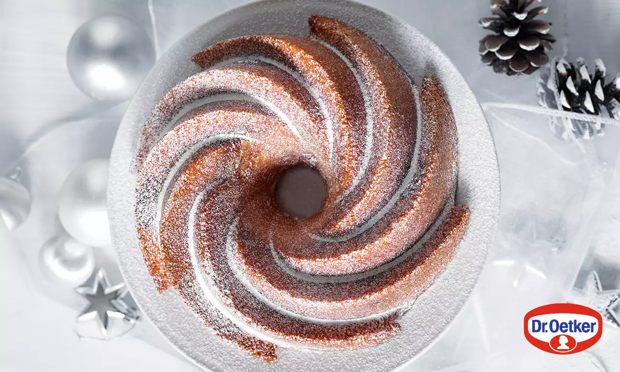 Vánoční bábovka s hnědou zlatavou kůrkou, spirálově tvarovaná, posypaná bílým moučkovým cukrem na stříbrném talíři mezi vánočními ozdobami