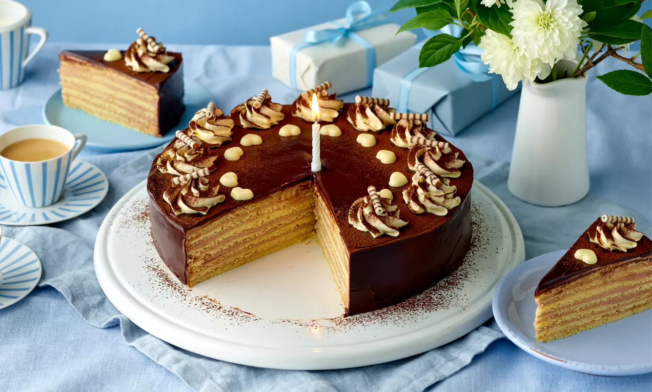 Prinzregententorte mit dunkler Schokoladenglasur, heller Schichtoptik und cremigen Verzierungen auf weißer Tortenplatte angerichtet