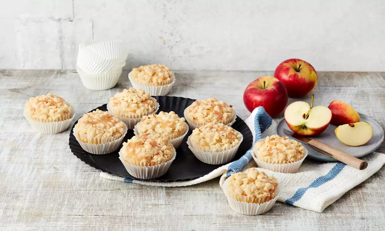 Apple streusel muffins with golden crumbly tops in white paper liners on a black plate, surrounded by fresh apples and sliced apple wedges on a cloth