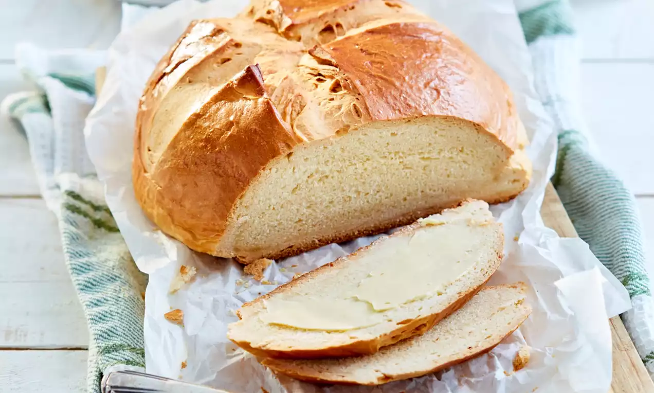 Pâine rotundă cu crustă aurie, ușor crestată, așezată pe hârtie de copt, alături de câteva felii proaspăt tăiate și un cuțit pentru unt