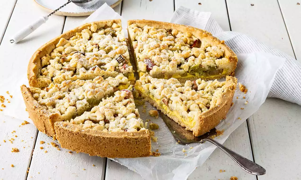 Gâteau rond avec une pâte dorée, garniture rhubarbe jaune entrevue sous des streusels croustillants saupoudrés de sucre sur papier blanc